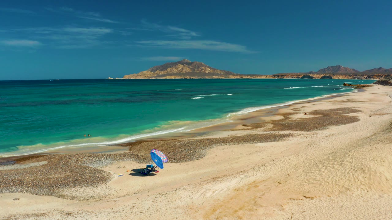 Turquoise shoreline with swimmers, blue umbrella, sand patches, and mountains.