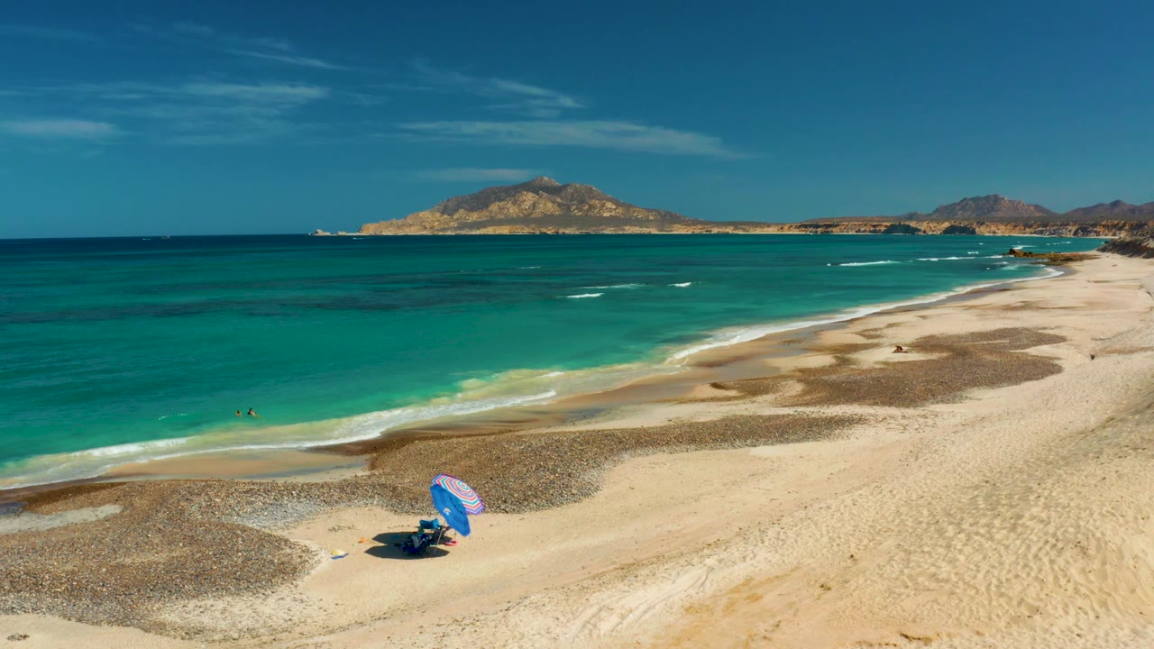 Cabo Pulmo beach two seconds in, with the umbrella shifted toward the lower center of the frame, swimmers in the surf zone, and a long pale shoreline running right.