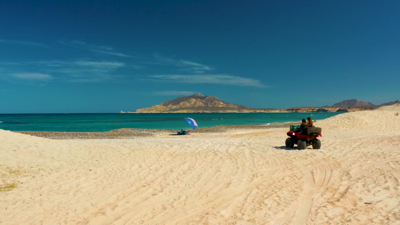 At the start of the Cabo Pulmo clip, a red ATV with two visible people sits on pale sand near a blue umbrella, with turquoise water and distant hills under a clear sky.