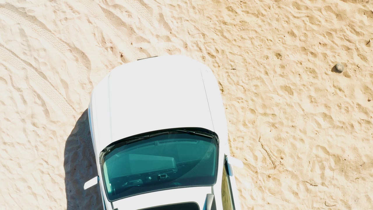 Overhead frame from Cabo Pulmo, Baja California Sur showing the upper half of a white car on pale tracked sand, with a dark side shadow and no visible people.