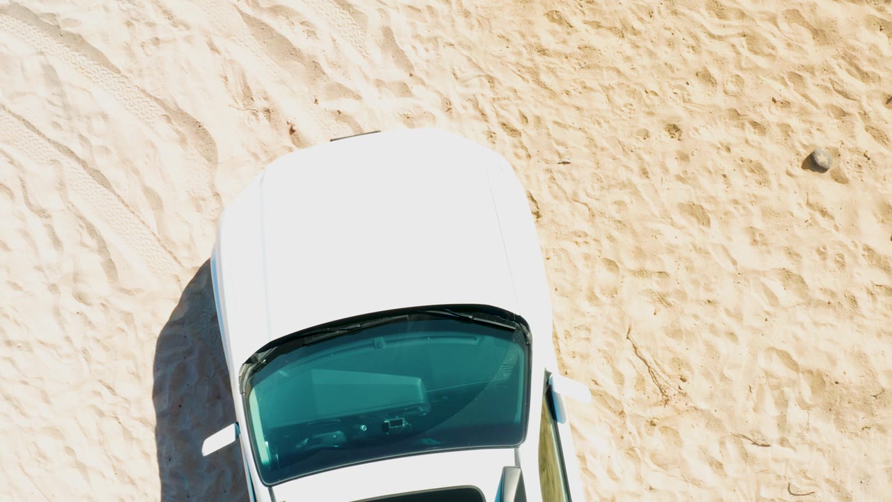 Top-down Cabo Pulmo frame showing the white car slightly farther along the sandy track, with strong sunlight, a left-side shadow, and no visible people.