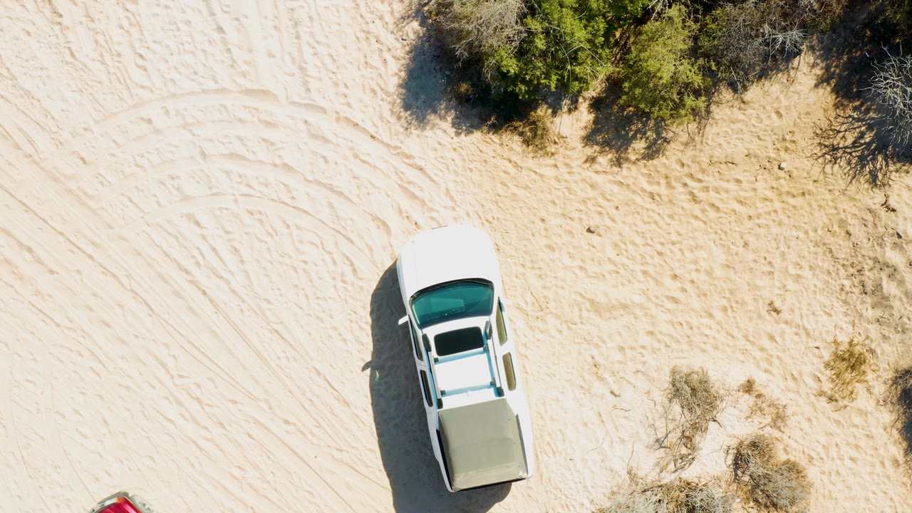 Overhead view of a white vehicle entering sandy terrain in Cabo Pulmo, Baja California Sur, with green bushes along the upper edge and no visible people.