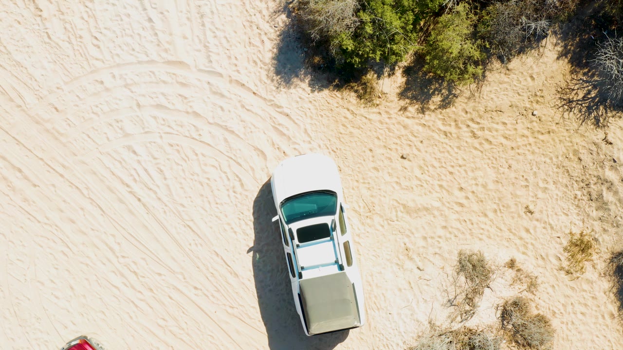 Overhead frame of the white vehicle slightly farther across pale sand in Cabo Pulmo, with tire tracks, scrub vegetation, and no visible people.