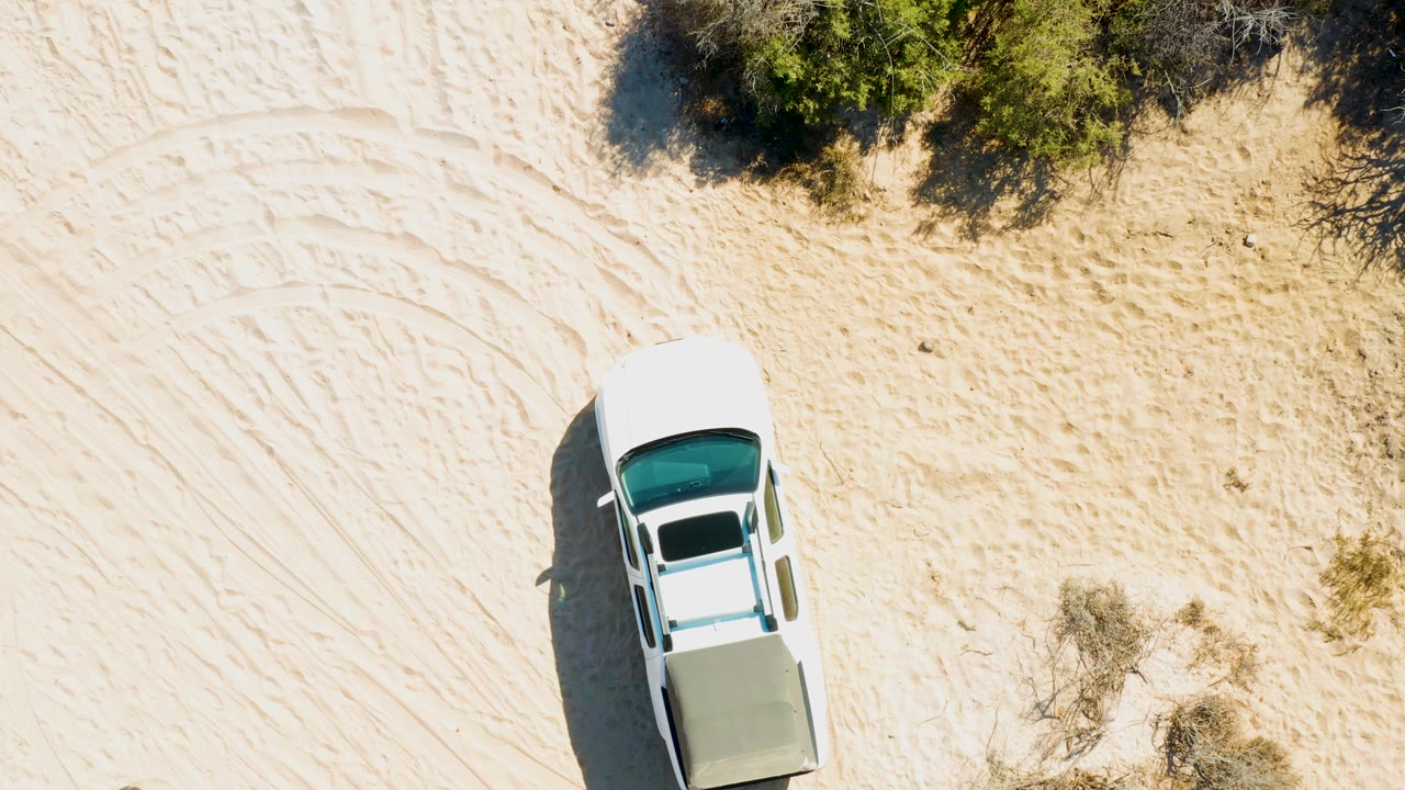 Top-down view of a white vehicle crossing open sand in Cabo Pulmo, Baja California Sur, with scattered brush and no visible people.