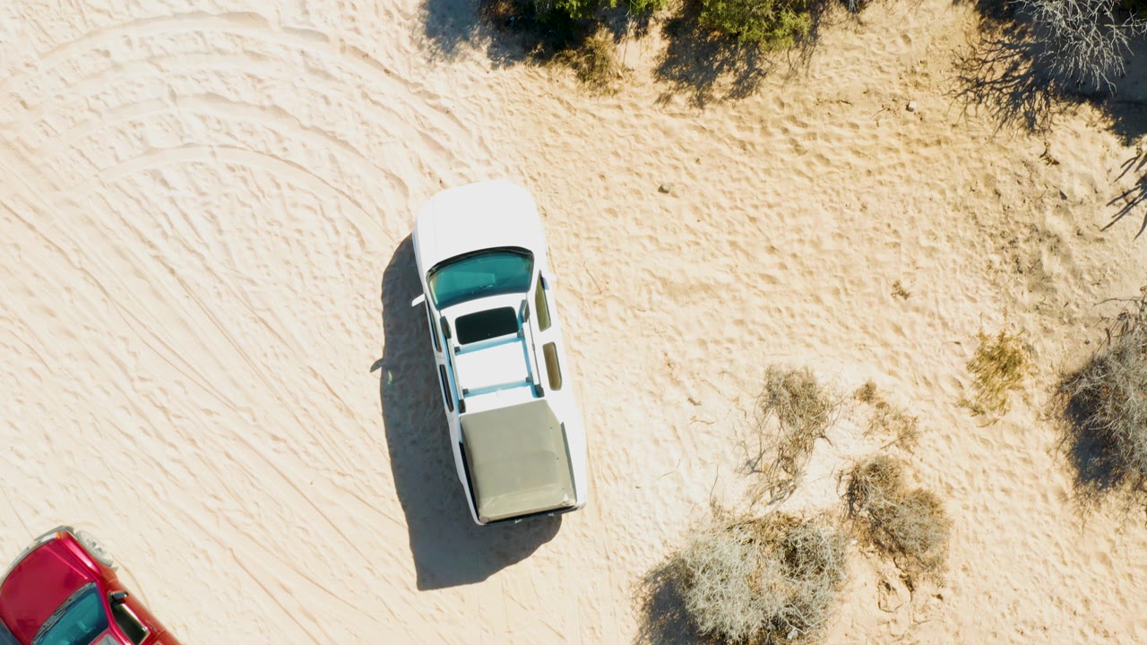 Overhead frame in Cabo Pulmo, Baja California Sur showing a white pickup on pale sandy terrain, scrub bushes at the top edge, a partial red car at lower left, and no visible people.