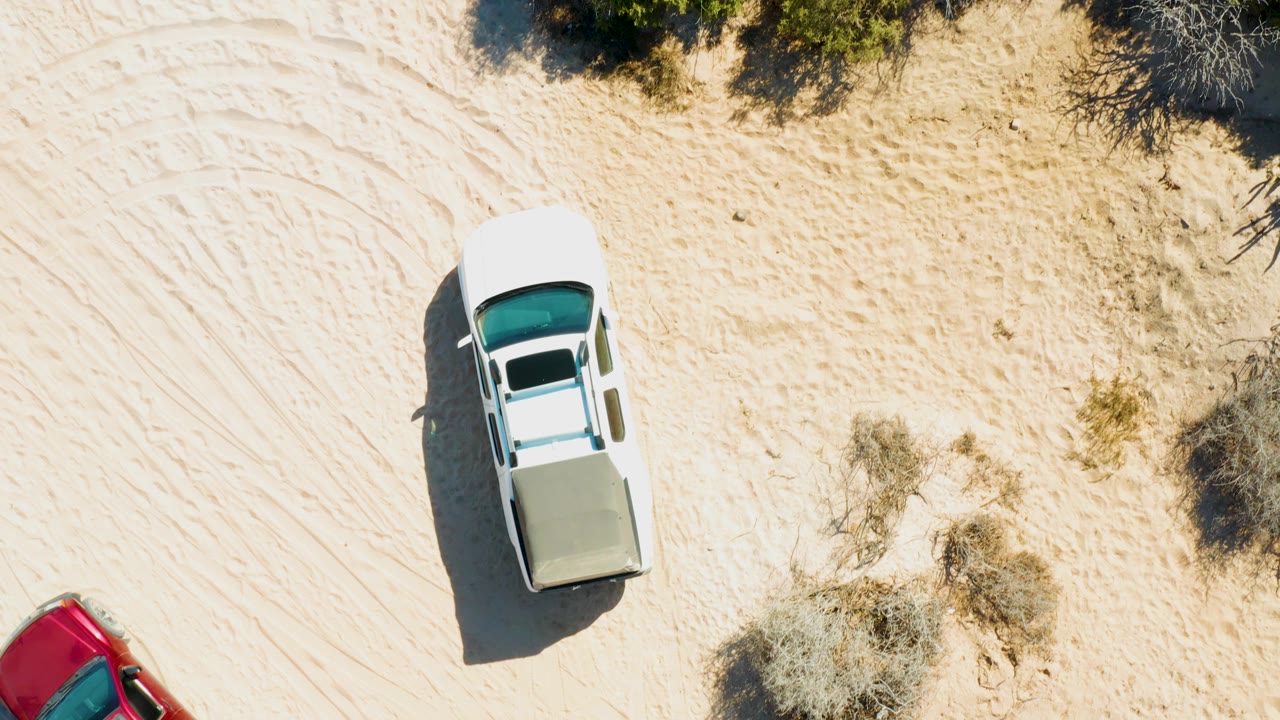 Overhead frame in Cabo Pulmo, Baja California Sur showing the white pickup farther across pale sand, curved tire tracks, scrub vegetation along the upper edge, and no visible people.