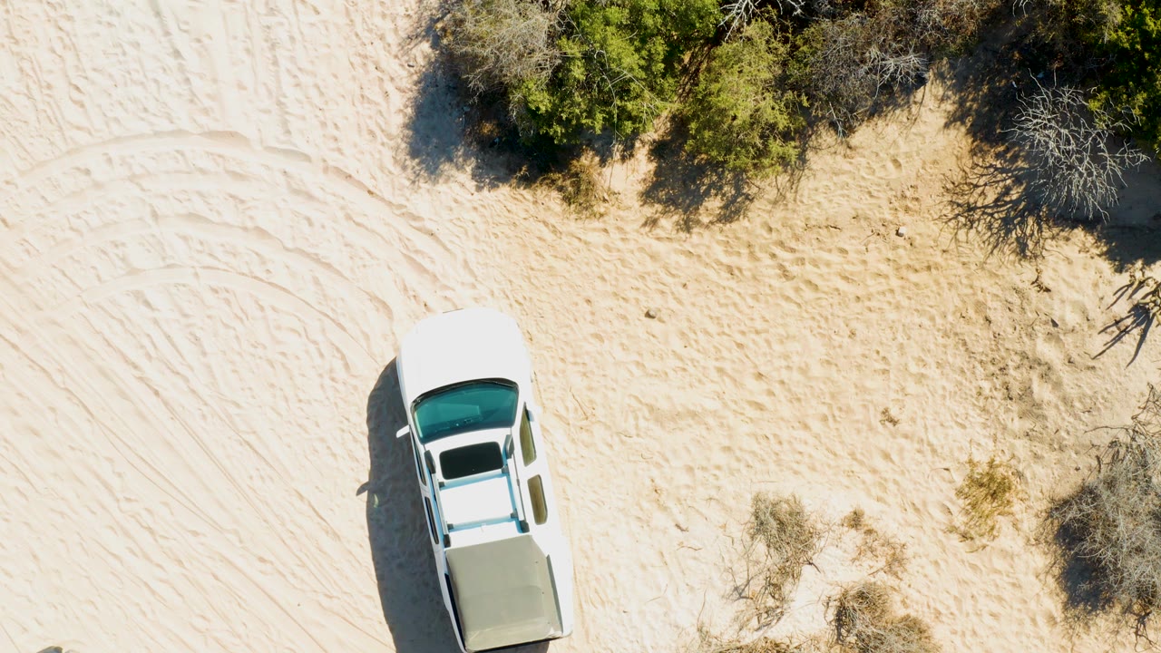 Overhead frame in Cabo Pulmo, Baja California Sur showing the white pickup lower in the composition, open sand with track marks, scrub at the top, and no visible people.