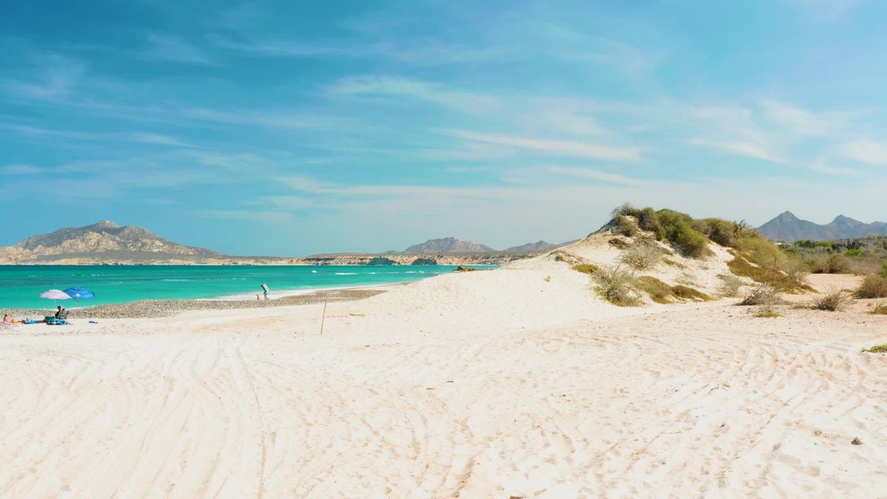 White sand beach with tire tracks, dunes, turquoise ocean, and distant mountains.
