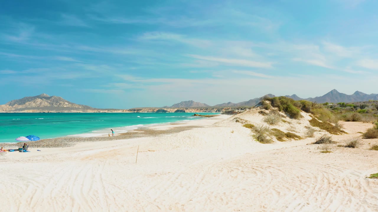 Beach curve with turquoise water, white sand, dunes, and small people near shore.