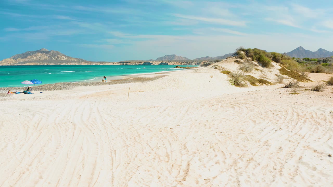 Sandy shore and turquoise water with people near a blue umbrella and dunes on the right.