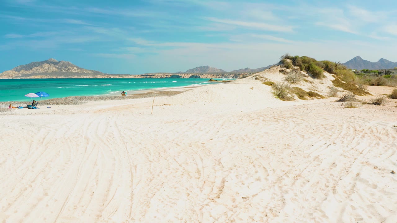 Wide Cabo Pulmo beach frame with the umbrella and people set far left, turquoise water running across the midground, a sandy dune mound at right, and mountain shapes on the horizon.