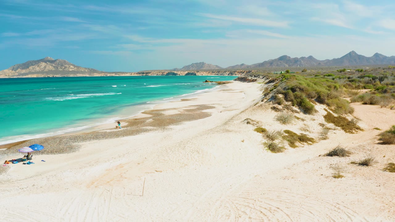 Wide drone-like view of a Cabo Pulmo sandy beach with turquoise water at left, dune grass at right, a small swimmer near shore, a blue umbrella low on the sand, and mountains on the horizon.