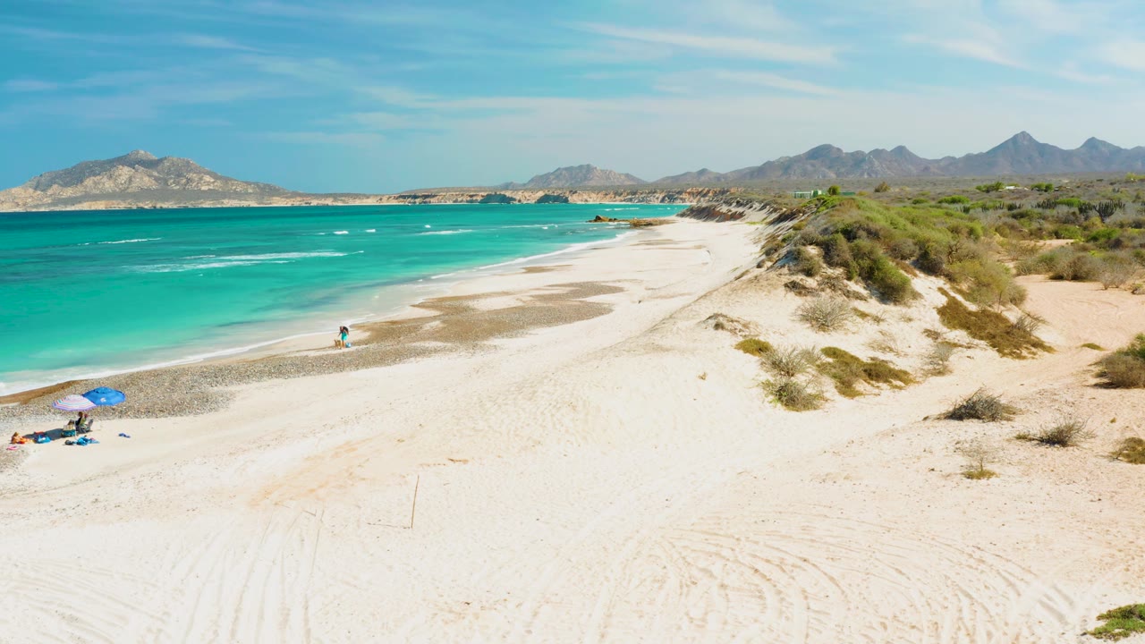 A wide Cabo Pulmo shoreline view shows pale sand in the foreground, turquoise ocean and a swimmer near the left, a blue umbrella with beach items below, dune shrubs to the right, and mountains under a clear sky.