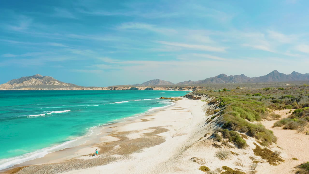 Wide elevated view in Cabo Pulmo showing turquoise water on the left, a pale sandy shore below, scrubby dunes to the right, distant mountains, and small beachgoers near the waterline.