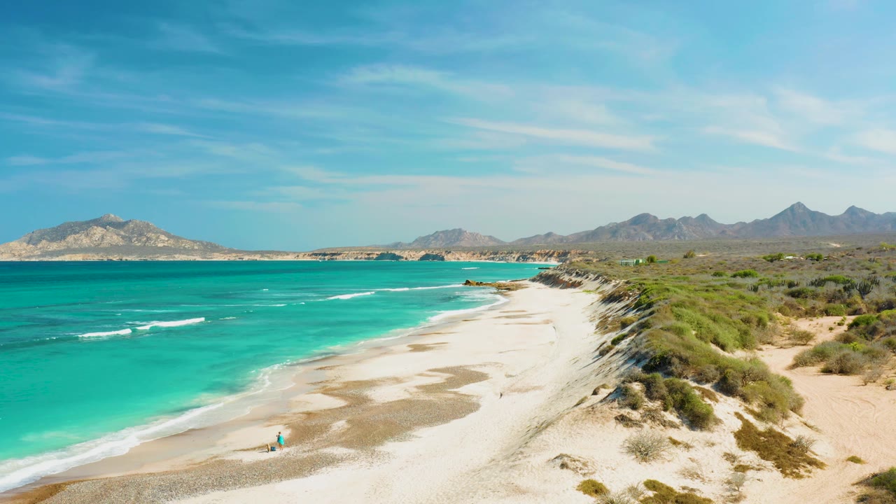 Turquoise waves beside a sandy shore with dunes, distant mountains, and small people on the beach.
