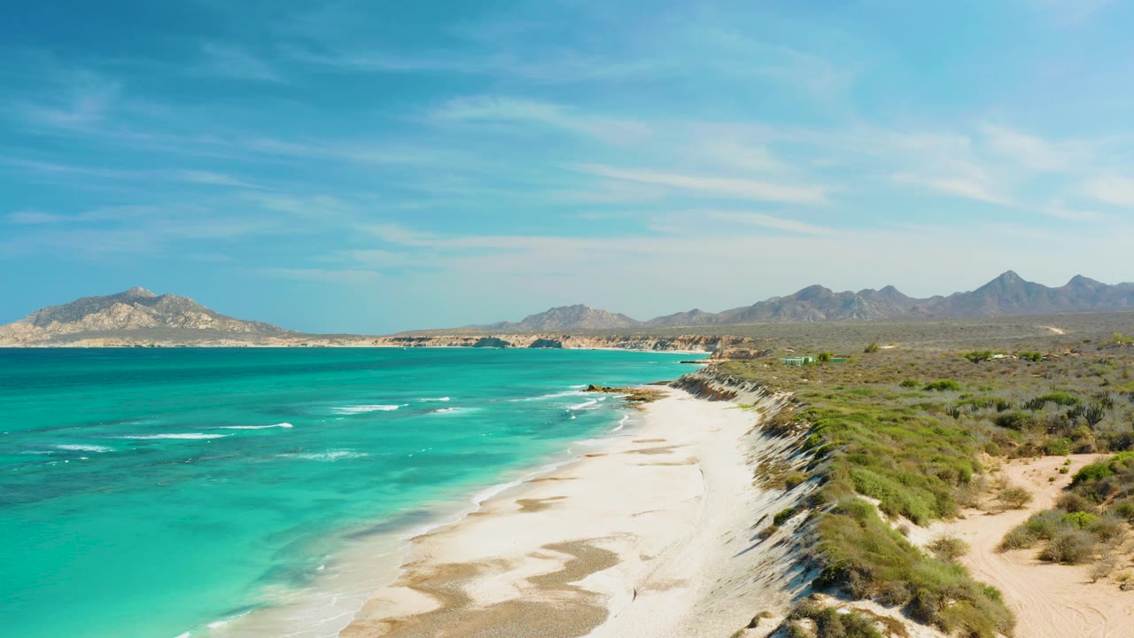 Elevated view of Cabo Pulmo coastline with turquoise ocean on the left, a pale sandy beach and scrub dunes on the right, distant mountains, and no visible people.