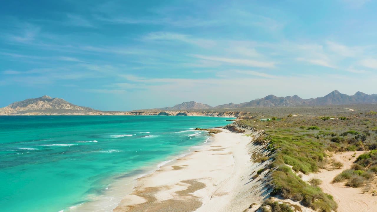 Wide coastal view with turquoise sea, pale beach, dunes, and mountains.