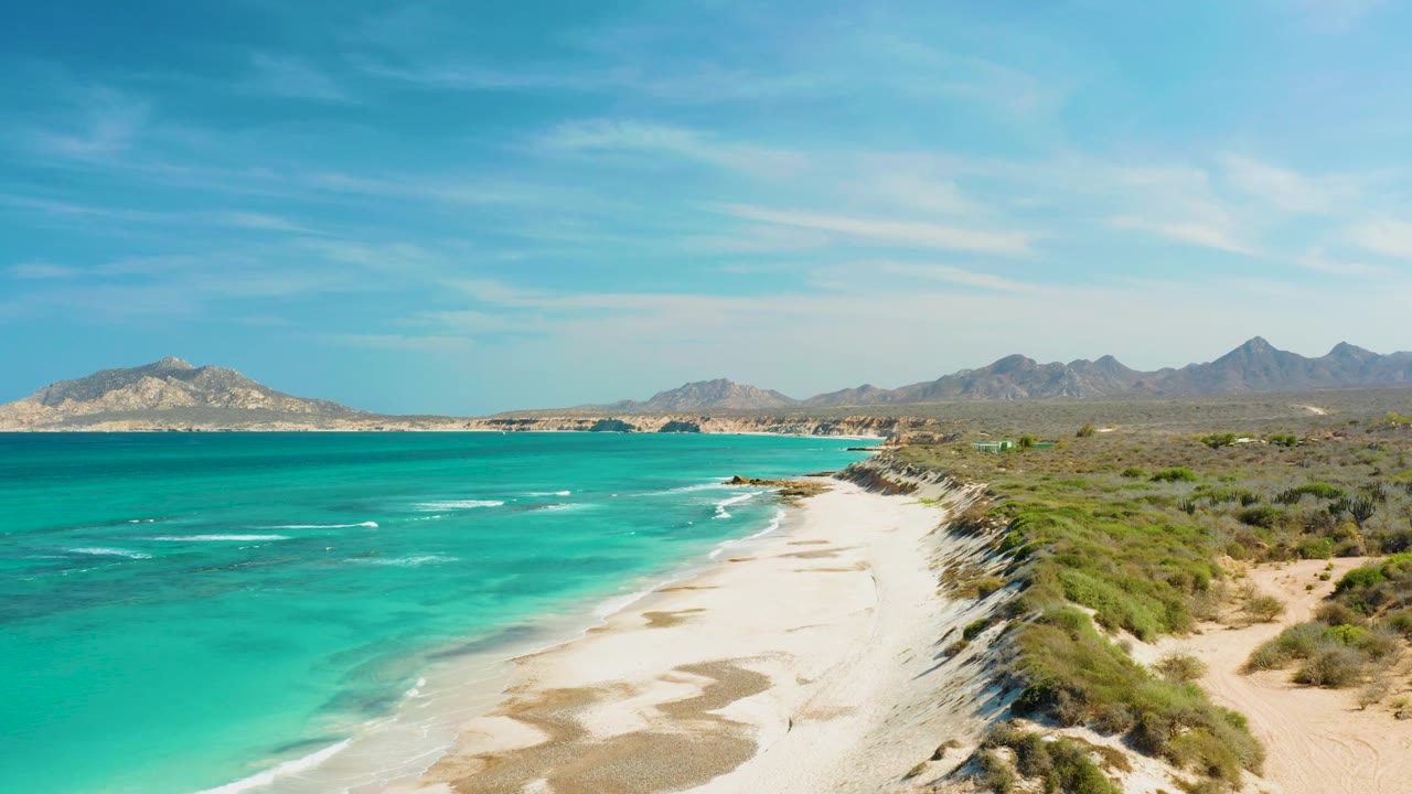 Cabo Pulmo shoreline curves through frame with turquoise water, broken white wave marks, pale sandbars, dune brush, distant mountains, and no visible people.