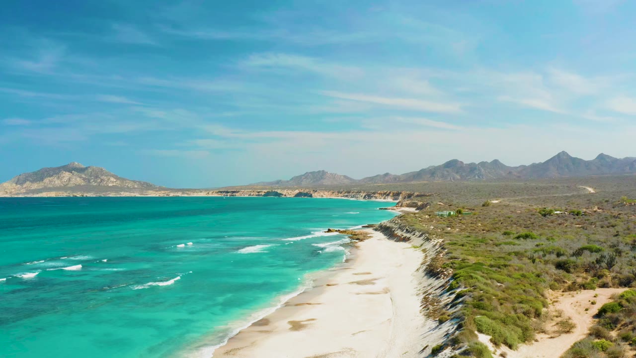 Aerial view of Cabo Pulmo coastline with turquoise water on the left, pale sandy beach below, scrub-covered cliffs to the right, distant mountains, clear blue sky, and no visible people.