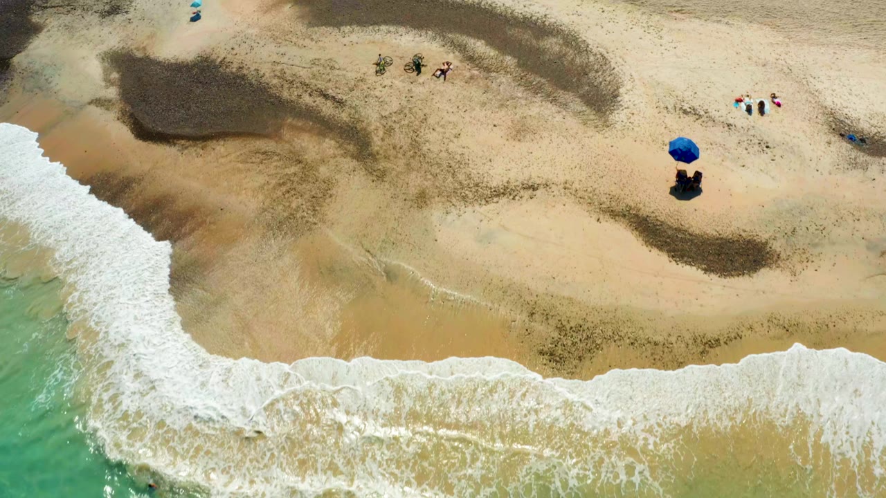 Aerial shoreline with white waves and small figures on sand.
