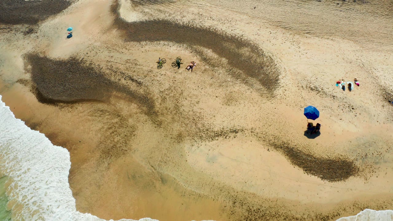 Overhead Cabo Pulmo beach view with the surf edge slipping toward the lower frame, a blue umbrella at right, bicycles near the upper sand, and people in small clusters.