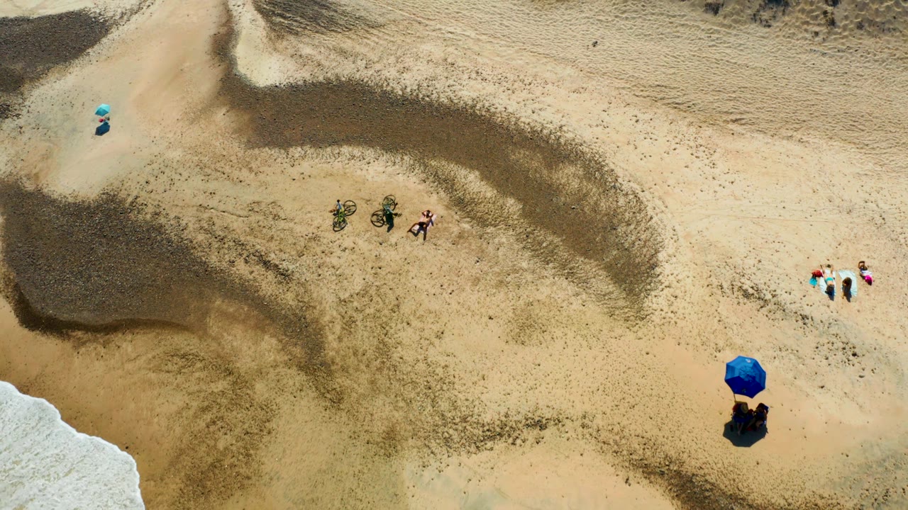 Beach sand with people, bicycles, umbrella, and wave foam at corner.