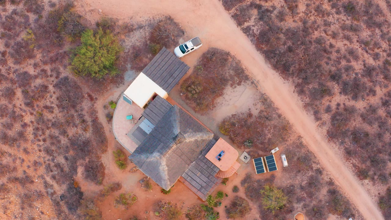 Overhead view in Cabo Pulmo, Baja California Sur, showing a desert house, pale dirt road, parked white car, solar panels, red-brown soil, sparse shrubs, and no visible people.