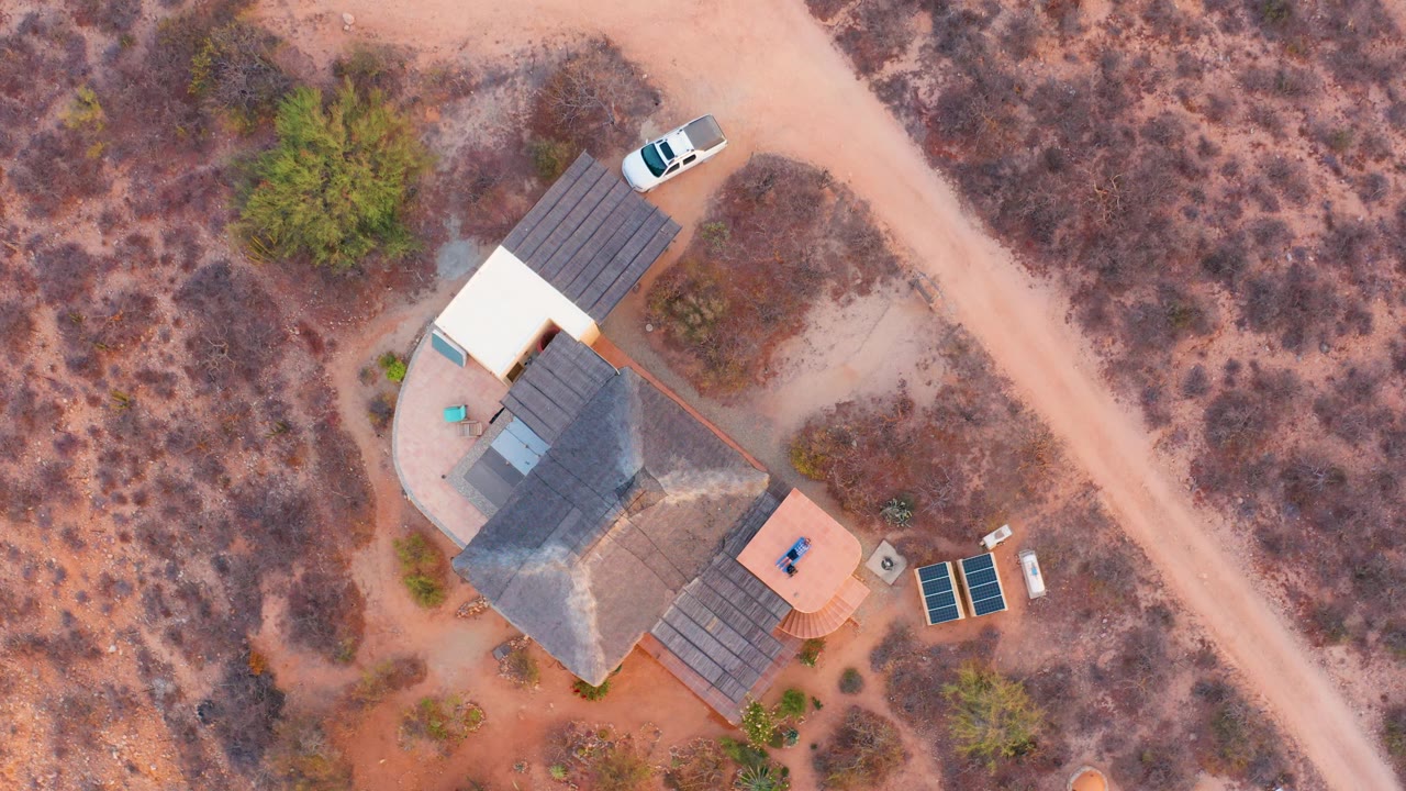 Top-down frame in Cabo Pulmo, Baja California Sur, showing the residence beside the diagonal dirt road, a white parked car near the roofline, solar panels below, and no visible people.