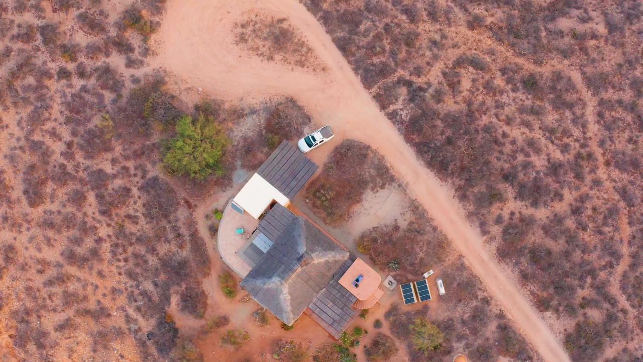 Overhead drone frame in Cabo Pulmo, Baja California Sur showing a small desert house compound, pale dirt road, nearby white car, scrub vegetation, and no visible people.