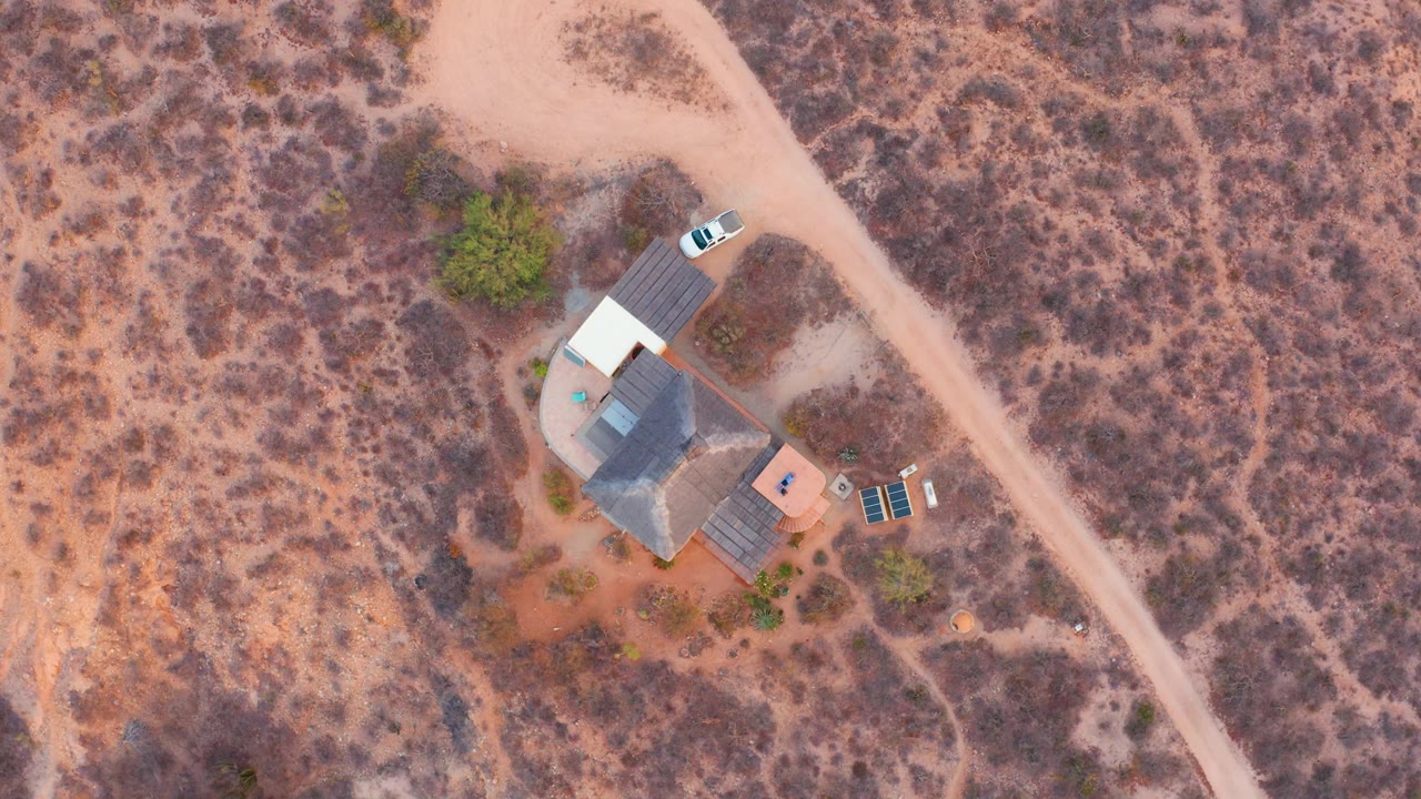 Overhead frame in Cabo Pulmo, Baja California Sur showing a small desert house, a white car near the dirt road, solar panels, dry brush, reddish soil, and no visible people.