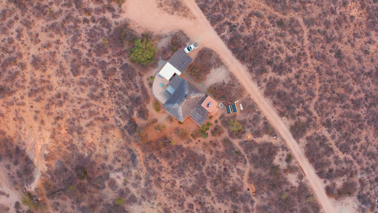 Overhead frame from Cabo Pulmo, Baja California Sur showing a small house compound, dry scrub, a pale dirt road, a parked white car, and no visible people.