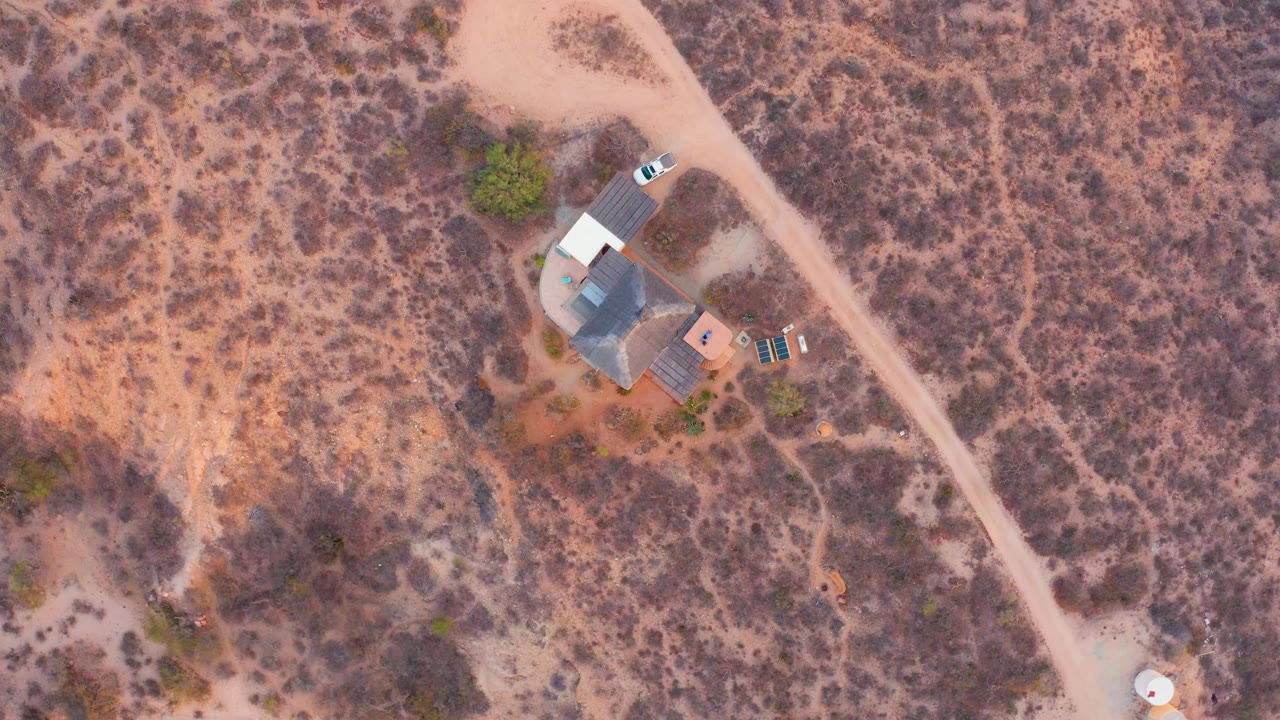 High overhead view in Cabo Pulmo, Baja California Sur, showing a white car close to a small house beside a pale dirt road, with dry scrubland around it and no visible people.