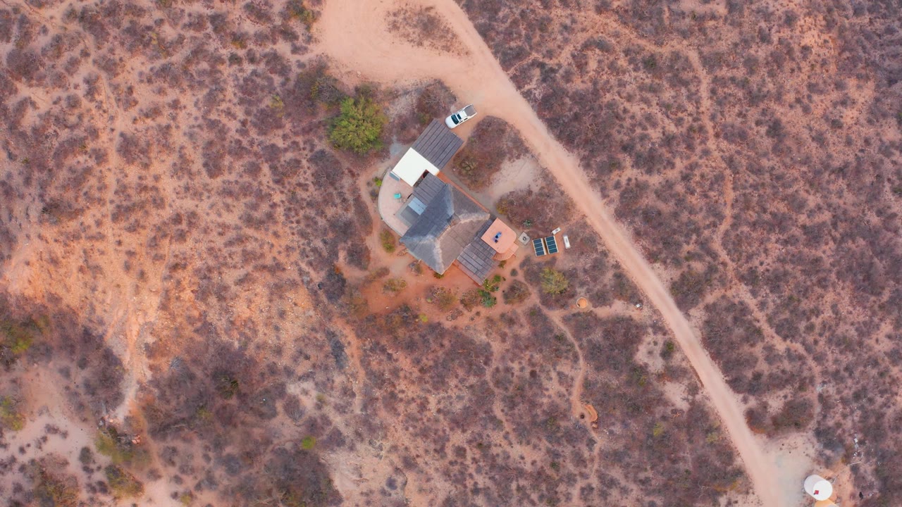 High drone view in Cabo Pulmo with the white car slightly farther up the road from the house, dry brush filling the background, and no visible people.