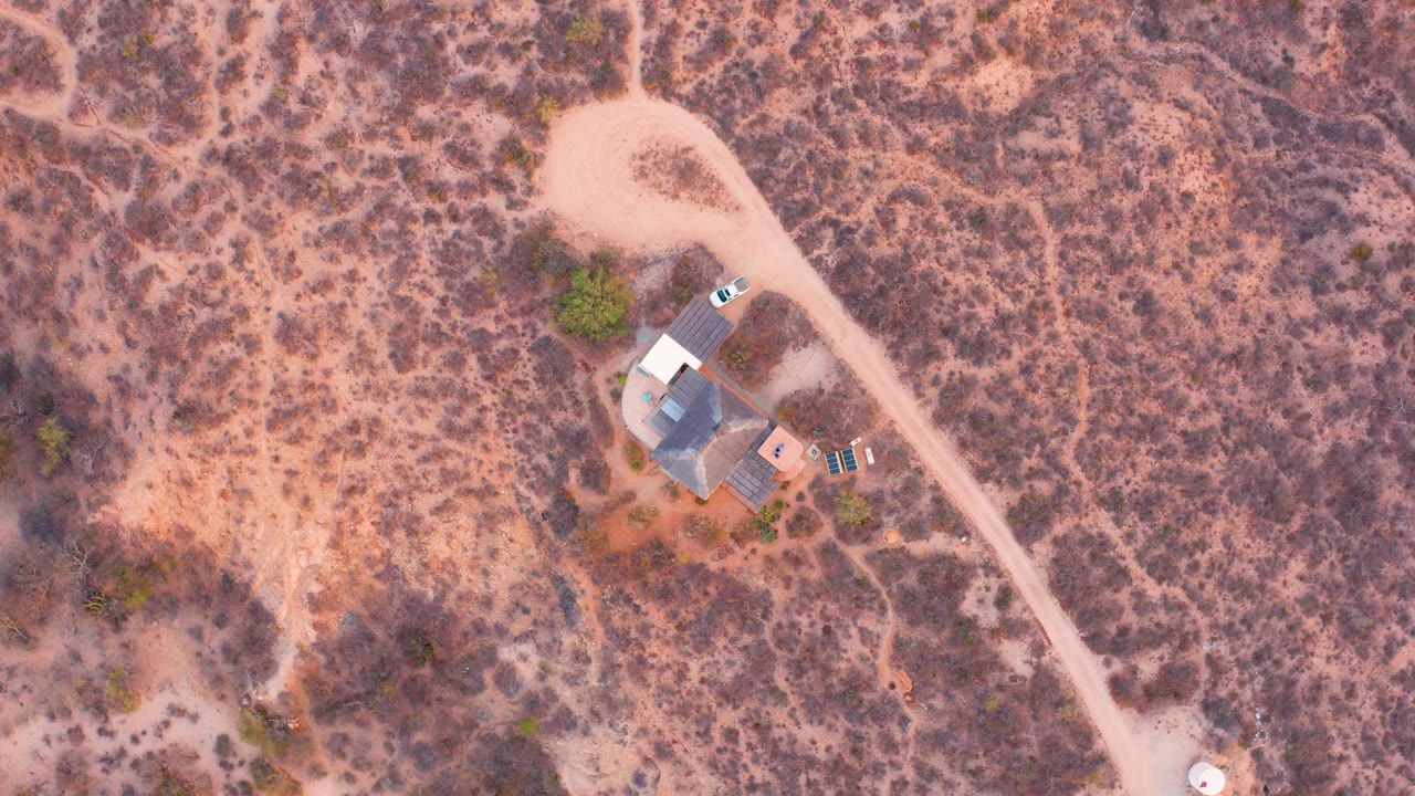 Near-vertical aerial view over Cabo Pulmo desert terrain with a small isolated house, a curving dirt road, parked vehicles, scrub vegetation, and no visible people.