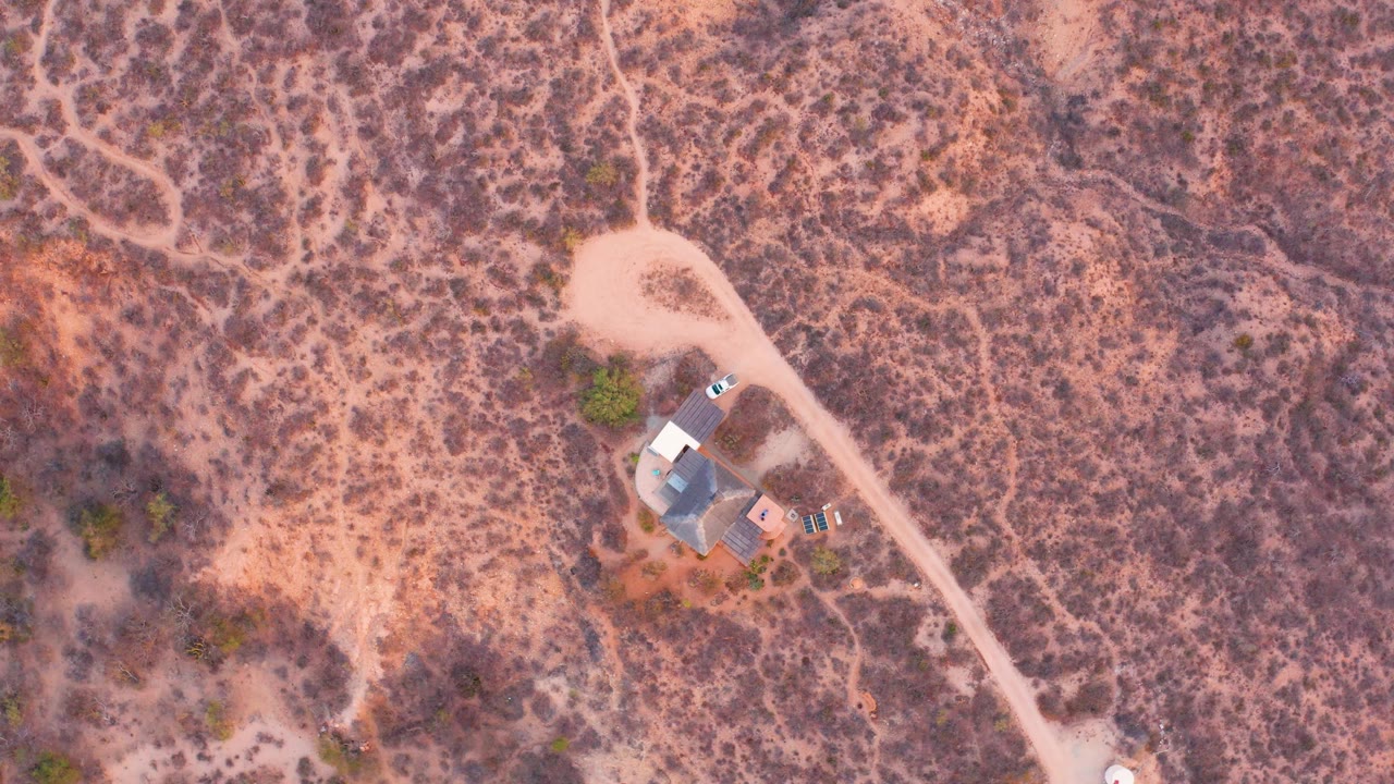 Overhead drone frame in Cabo Pulmo, Baja California Sur showing a pale dirt road beside a small house surrounded by dry scrubland, with no visible people.