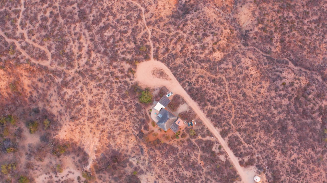 High drone view in Cabo Pulmo, Baja California Sur showing the remote house near a curved dirt road, mottled desert vegetation, pink-brown soil, and no visible people.