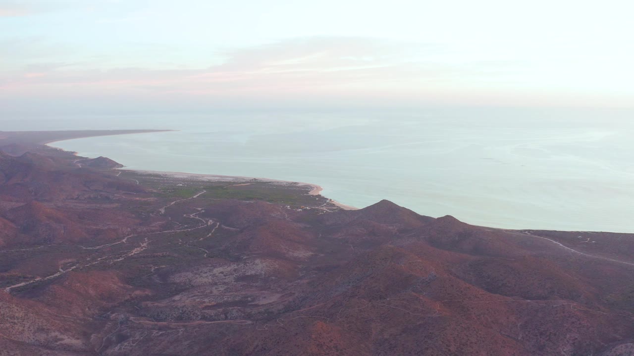 At 0:00, an aerial view over Cabo Pulmo shows red rocky mountain slopes leading to a pale beach and calm ocean under soft clouds, with no visible people.
