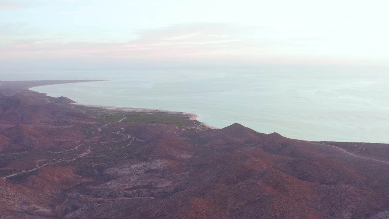 At 0:01, the Cabo Pulmo coastline fills the frame with reddish hills in front, a narrow beach at left, and open water stretching to the horizon, with no visible people.