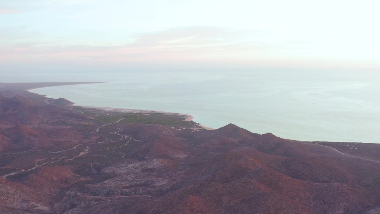 At 0:03, the aerial view over Cabo Pulmo shows the shoreline running along the left side of the water, red hills below, and a hazy horizon line, with no visible people.