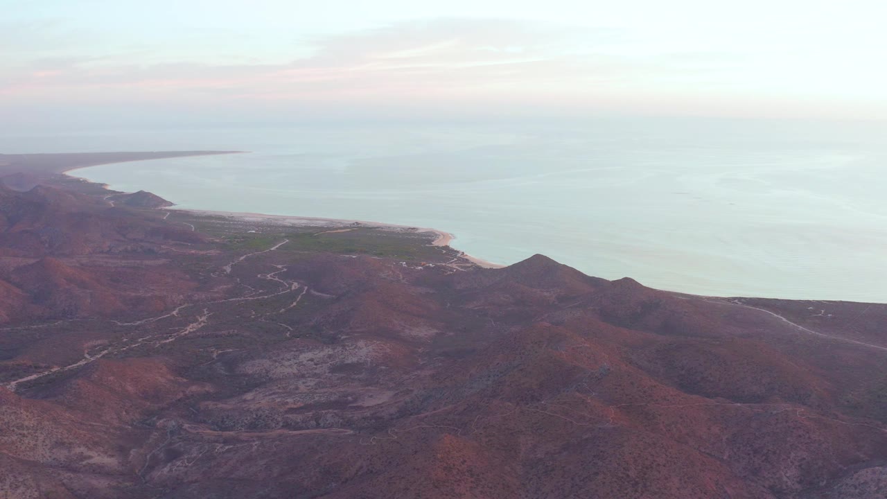 Cabo Pulmo aerial frame showing the curved coastline farther left, red-brown ridges across the lower frame, pale calm water in the background, and no visible people.