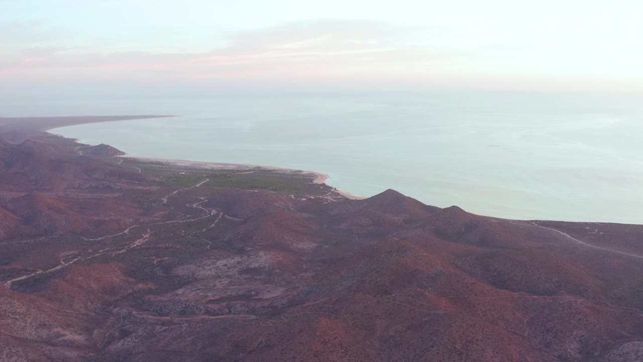 Mid-clip Cabo Pulmo aerial view with foreground ridges, faint winding tracks, a rounded shoreline, calm sea, pink-white sky, and no visible people.