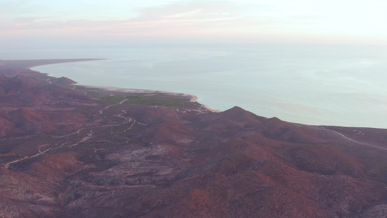 Wide Cabo Pulmo aerial view with mountain ridges in the foreground, a curved shoreline at left, and calm ocean under soft cloud color, with no visible people.