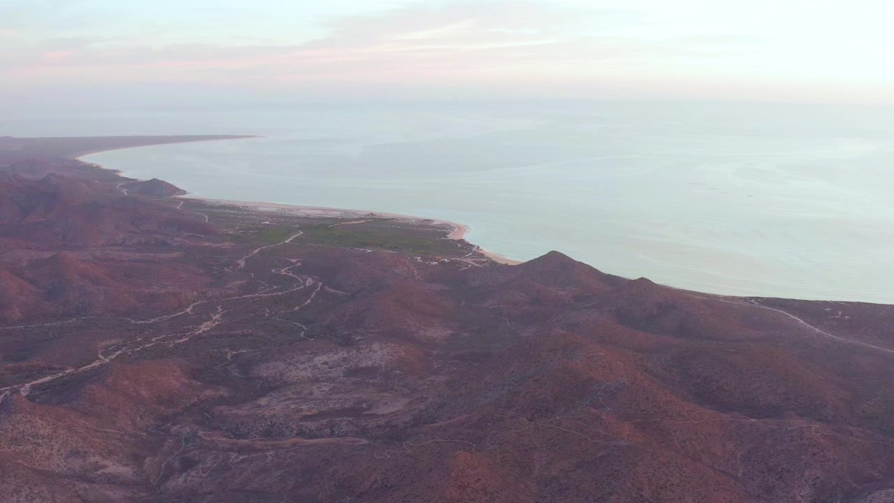 Cabo Pulmo aerial frame with dry ridges and winding tracks below, a small green area near the shore, pale sand along the waterline, open ocean, and no visible people.