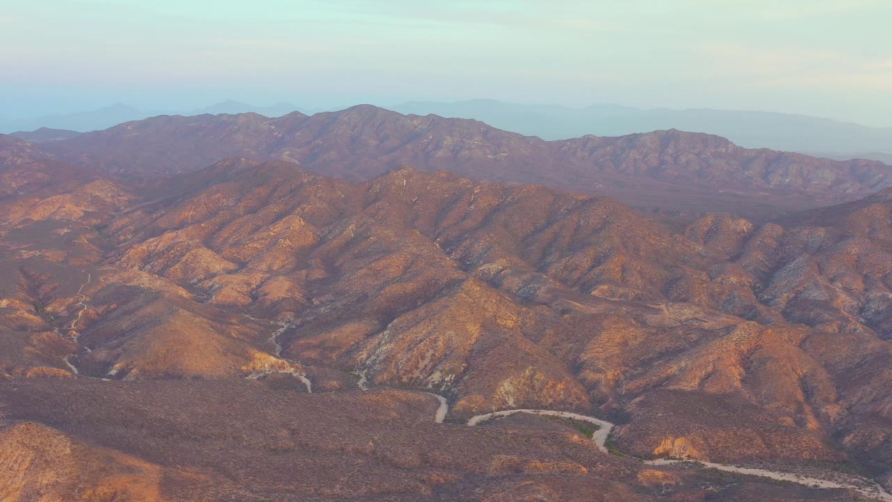 Opening aerial frame over Cabo Pulmo, Baja California Sur, showing dry brown ridges, a pale winding road in the lower valley, hazy distant mountains, and no visible people.