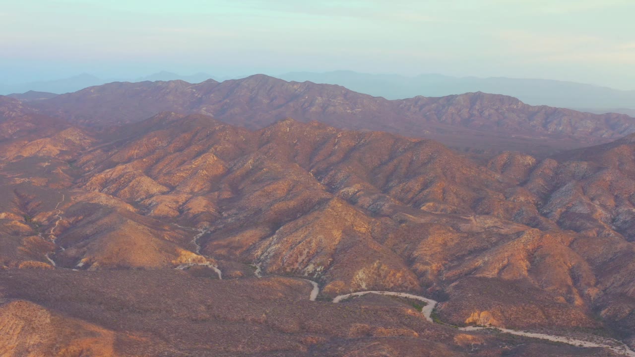 Aerial view over Cabo Pulmo, Baja California Sur, with the road bends visible near the bottom of the frame, layered brown hills across the center, distant ridgelines, and no visible people.