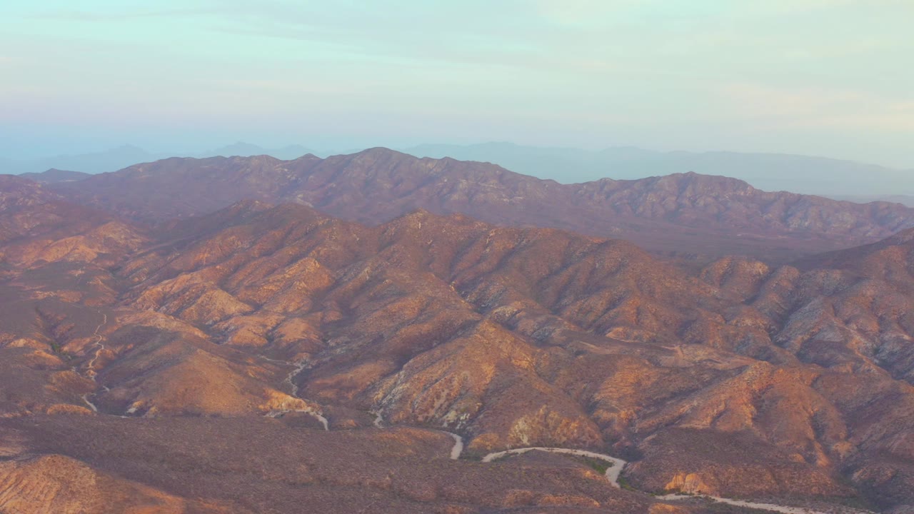 Aerial frame of brown ridges in Cabo Pulmo, Baja California Sur, with the winding road more visible across the lower valley and no visible people.