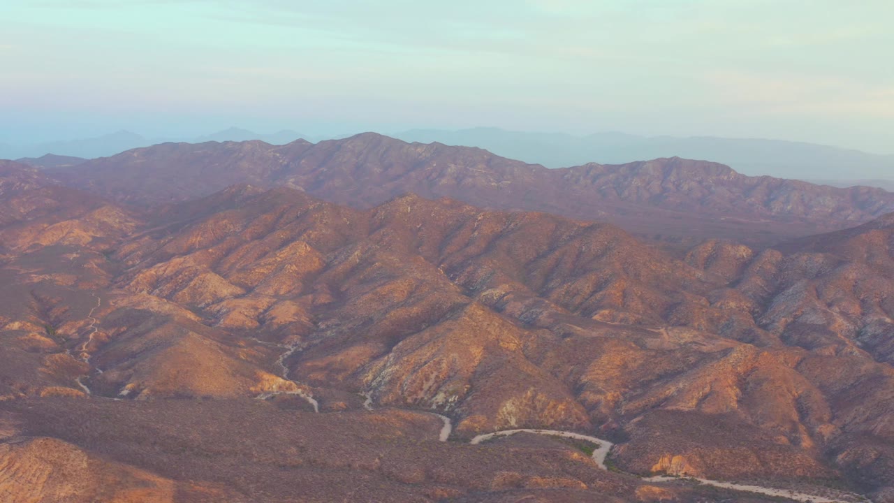 Mid-clip aerial view of Cabo Pulmo, Baja California Sur mountains, where shadowed ridges contrast with warm brown slopes and the winding road sits near the bottom edge, with no visible people.