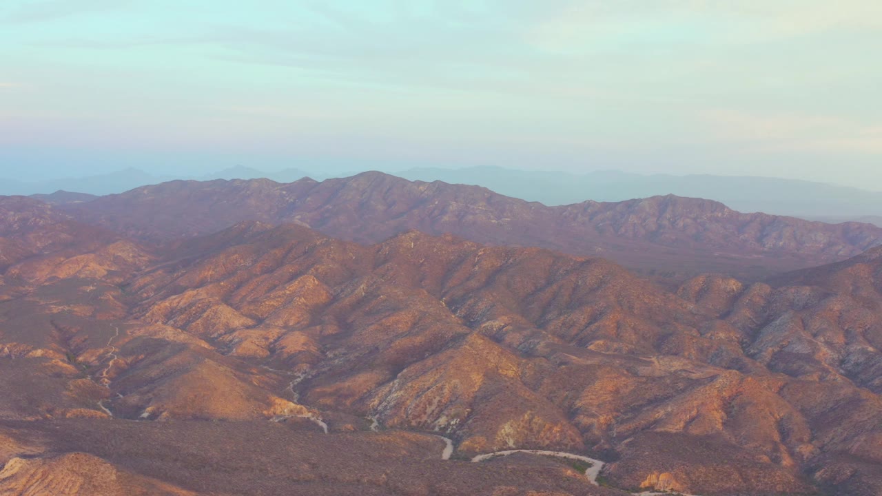 Aerial view in Cabo Pulmo with sunlit orange-brown hills, a curving road near the bottom of frame, and no visible people.