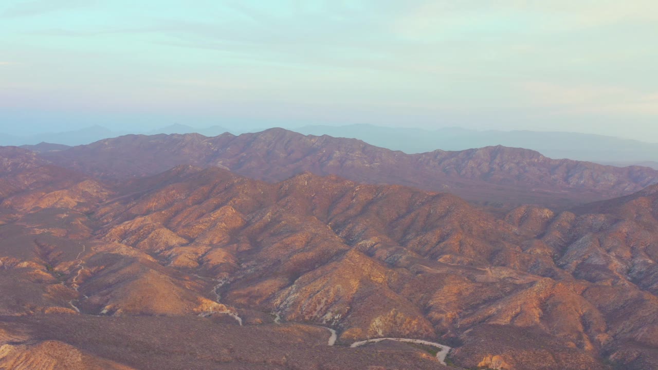 Mid-clip aerial frame over Cabo Pulmo hills, with warm light on dry ridges, the road curving across the foreground valley, and no visible people.