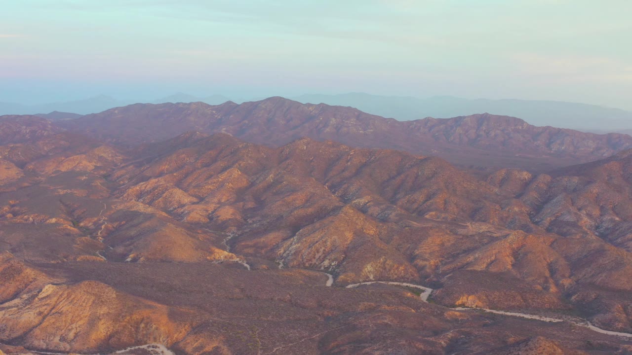 Aerial frame over Cabo Pulmo, Baja California Sur, showing sunlit brown ridges, a pale winding road across the valley floor, distant mountains, and no visible people.